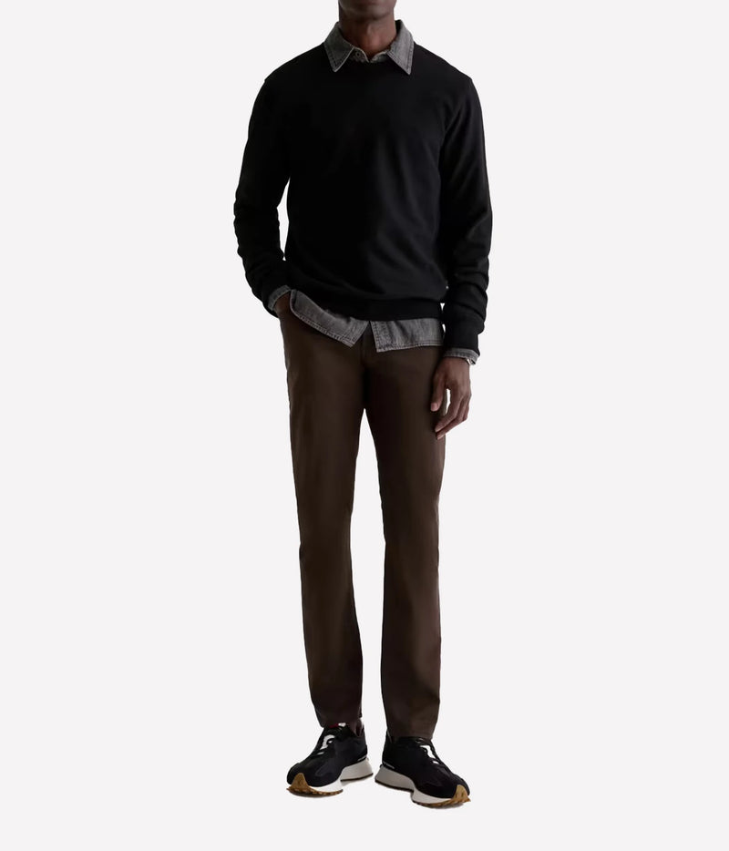 A man wearing a black crewneck sweater over a gray collared shirt, paired with brown trousers and black sneakers, shown against a plain light gray background.