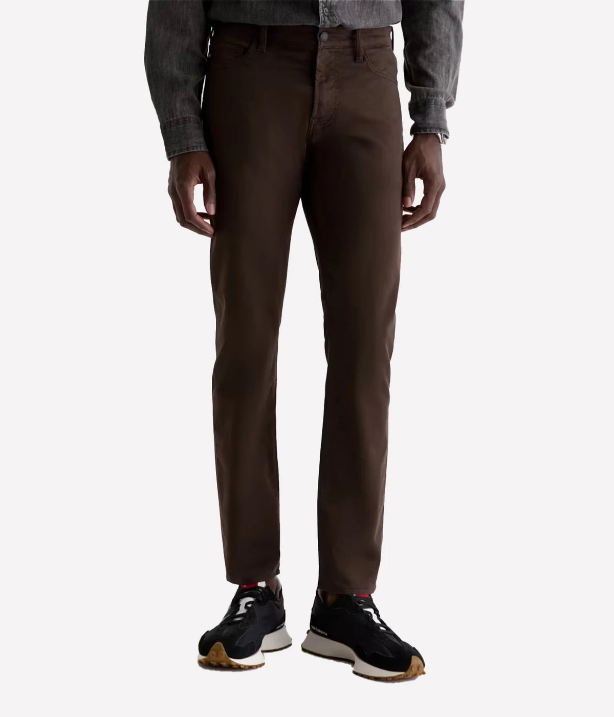 A person wearing slim-fit brown trousers, a gray denim jacket, and black sneakers, shown from the waist down against a light gray studio background.