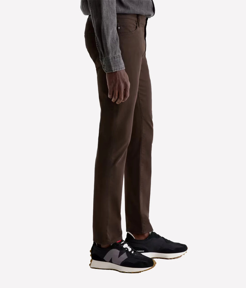 A side profile view of a person wearing slim-fit brown trousers, a gray long-sleeve shirt, and black New Balance sneakers against a plain light gray background.