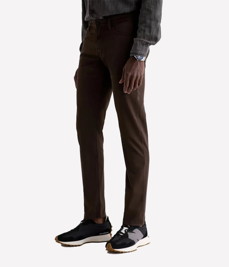A person wearing slim-fit dark brown trousers, a dark gray long-sleeve shirt, and black sneakers, shown from the waist down against a light gray studio background.