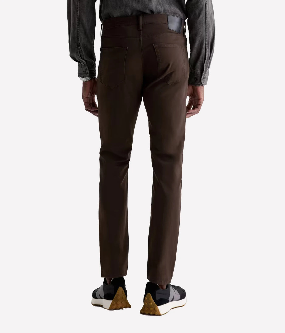 Rear view of a person wearing dark brown slim-fit trousers, a gray long-sleeve shirt, and black and gold sneakers against a light gray background.