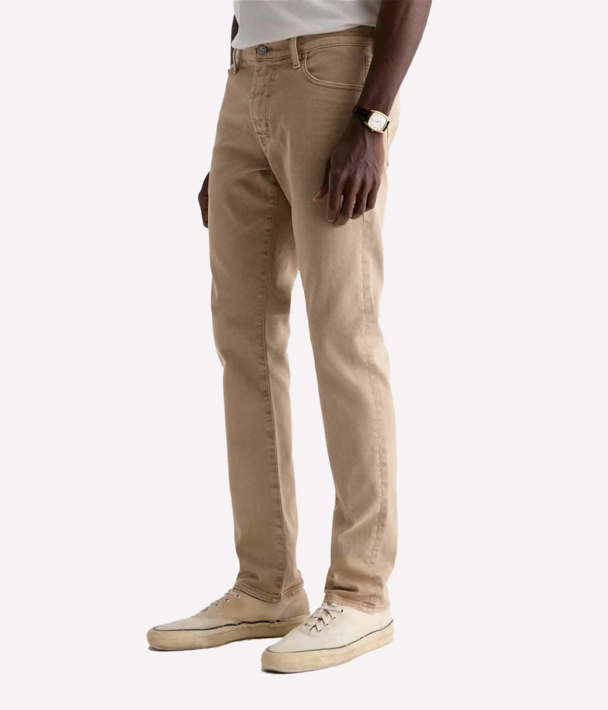 A person wearing slim-fit beige jeans paired with cream sneakers and a gold watch, shown from the waist down against a light gray studio background.