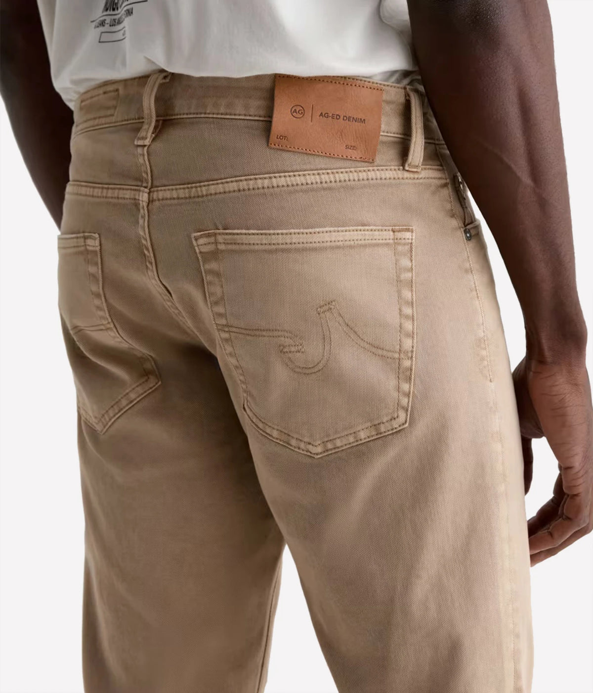 Close-up back view of a person wearing tan denim jeans, showing the waistband, leather brand patch, back pockets, and denim stitching.