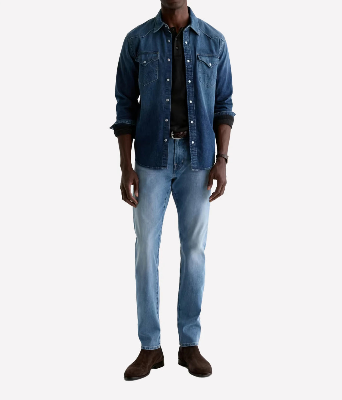 A man wearing a dark blue denim button-up shirt over a black collared shirt, paired with light blue jeans and dark brown suede boots, shown against a plain light gray background.