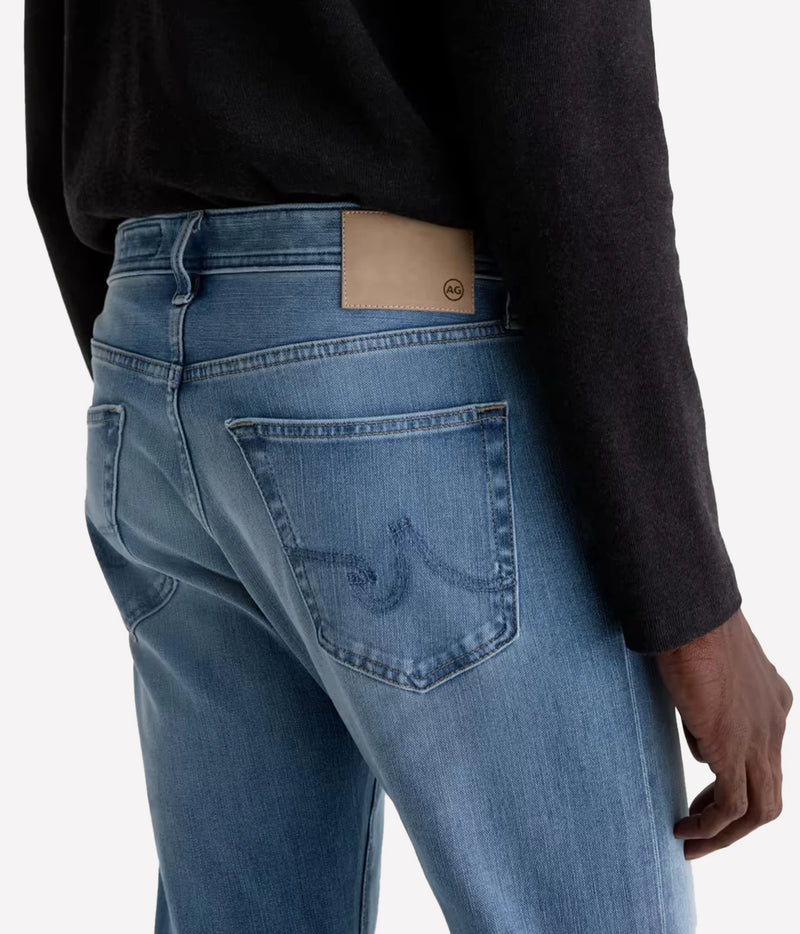 Close-up back view of a person wearing light blue denim jeans with a black long-sleeve top, showing the back pocket and waistband patch.
