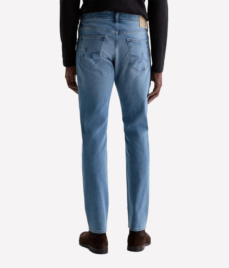 Rear view of a person wearing light blue slim-fit jeans with a black long-sleeve top and brown shoes, shown against a plain light gray studio background.