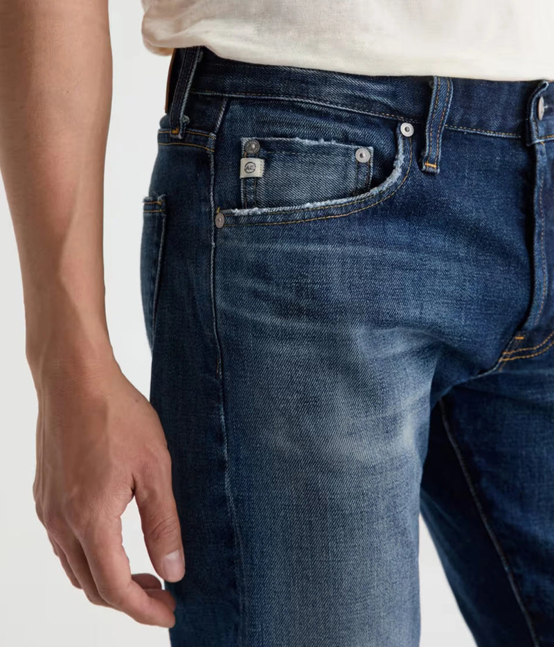 Close-up of a person wearing dark blue denim jeans with a light-colored t-shirt, showing the back pocket and waistband details.