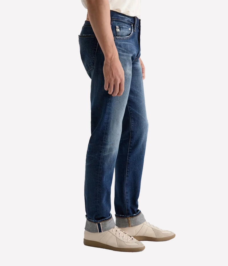 A side profile view of a person wearing dark blue slim-fit jeans with a faded wash, paired with off-white sneakers, against a plain light gray background.