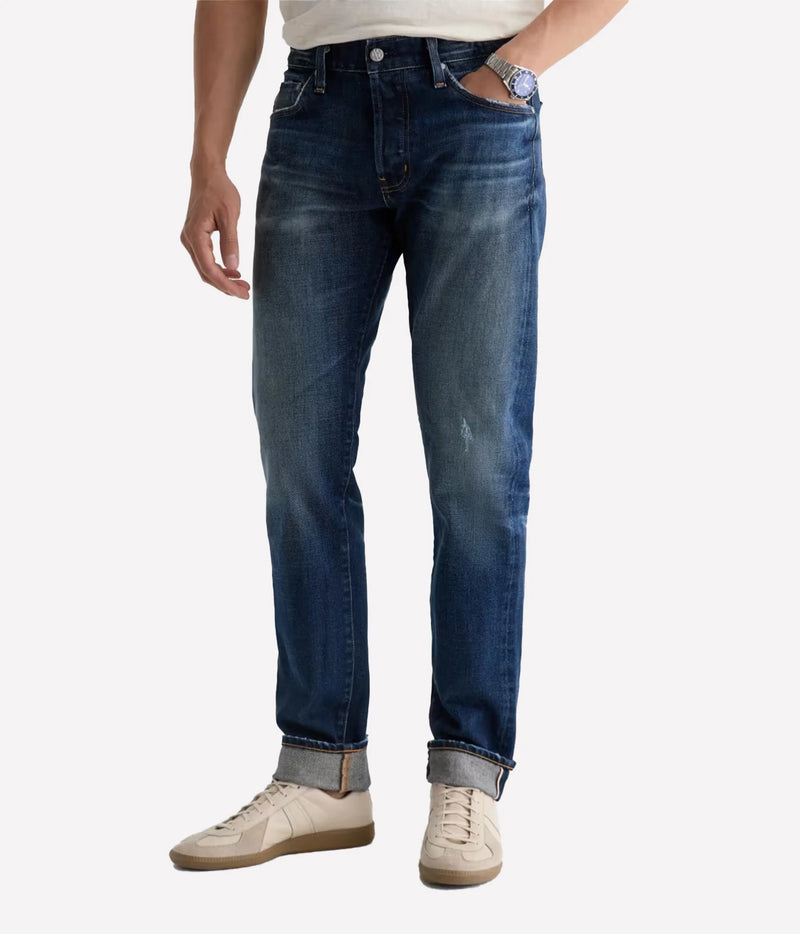 A person wearing a pair of medium-wash blue denim jeans with rolled cuffs, paired with off-white sneakers and a white shirt, shown from the waist down on a light gray background.