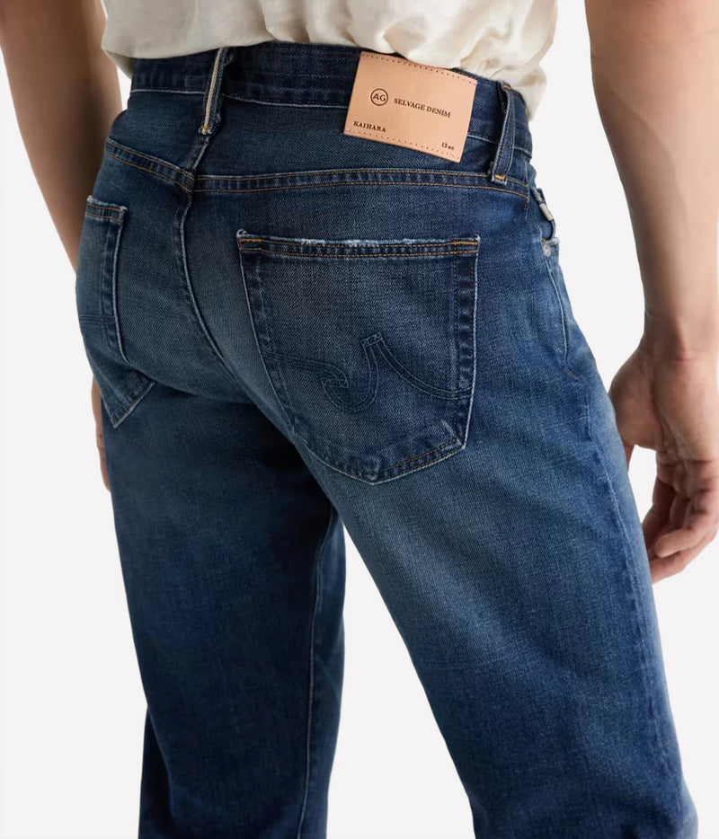 Close-up back view of a person wearing dark blue denim jeans with a tan leather brand patch at the waistband, photographed against a light gray studio background.