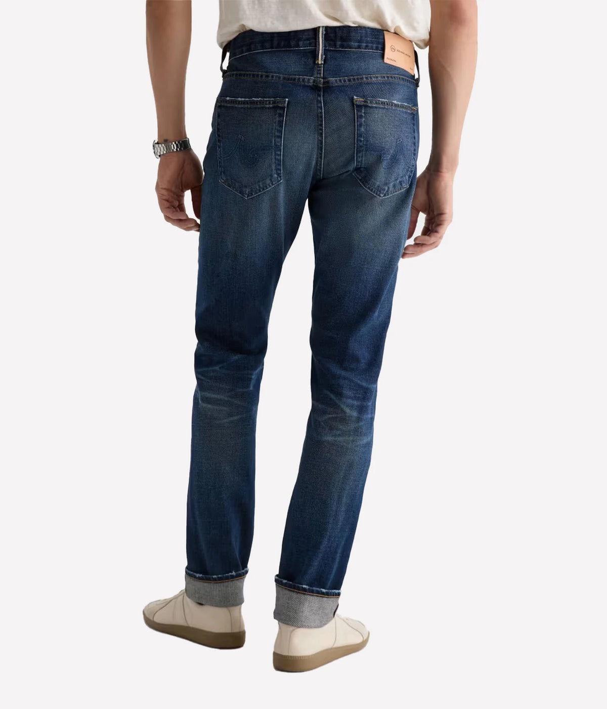 Rear view of a person wearing dark blue slim-fit jeans with a faded wash, paired with a light beige shirt and beige shoes, against a plain light gray background.