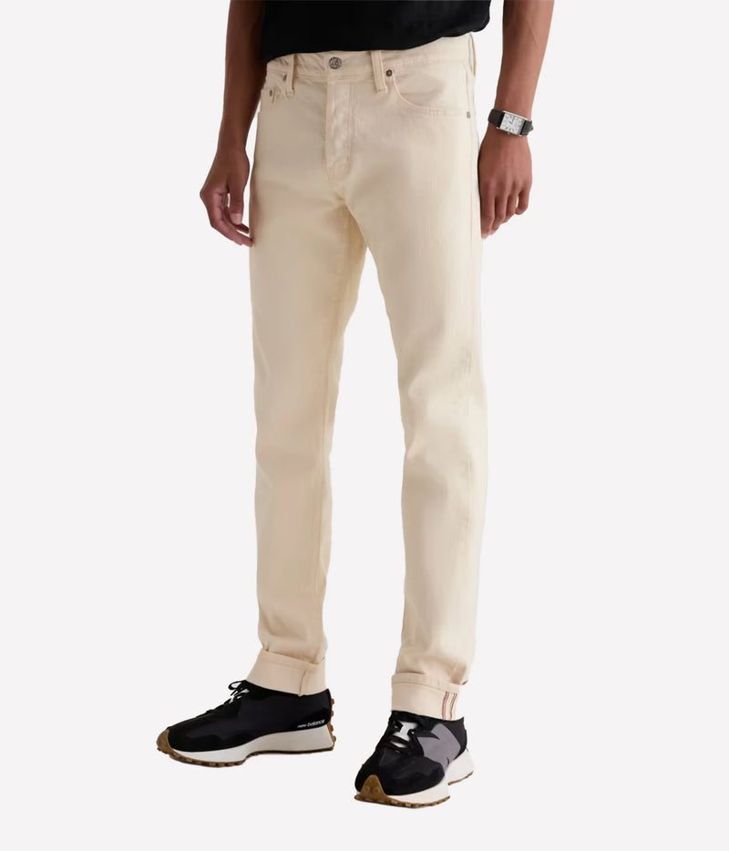 A person wearing slim-fit off-white denim jeans with black sneakers, shown from the waist down against a plain light gray studio background.