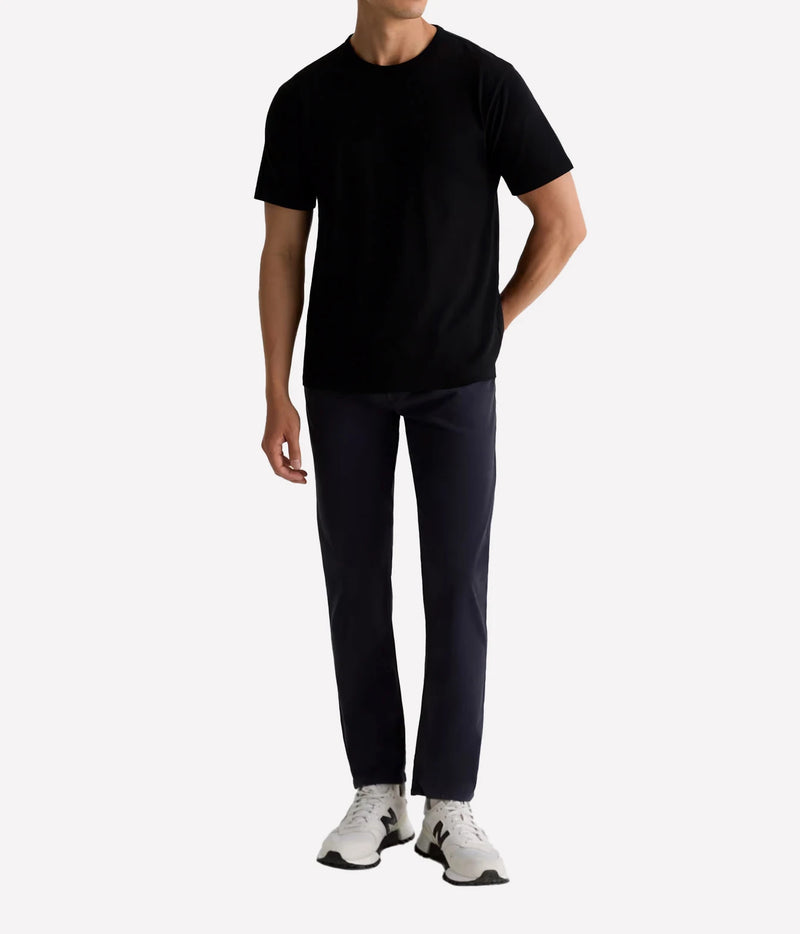 A man wearing a black short-sleeve crewneck t-shirt, dark navy pants, and white sneakers, standing against a light gray studio background.