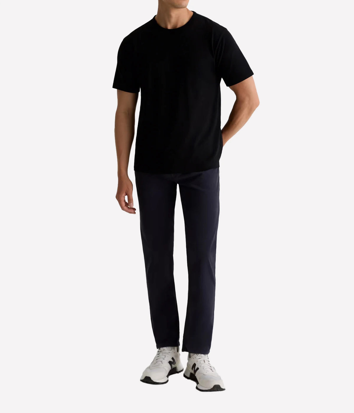 A man wearing a black short-sleeve crewneck t-shirt, dark navy pants, and white sneakers, standing against a light gray studio background.