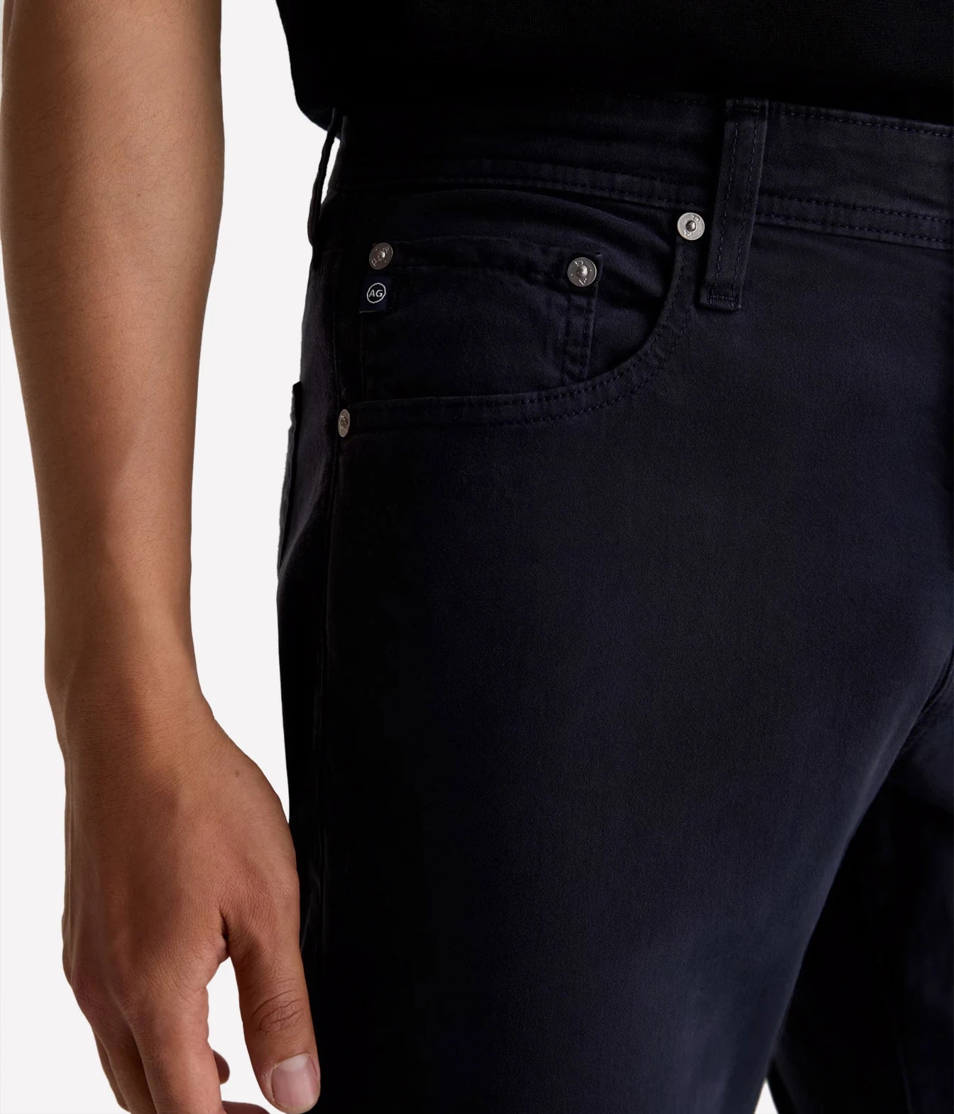 Close-up of a person wearing dark navy blue jeans, showing the back pocket area with silver-toned rivets and a metal button, against a plain light background.