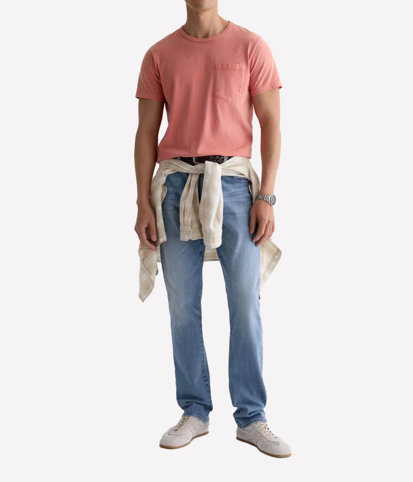 A model wearing a dusty pink crewneck pocket t-shirt, light-wash blue jeans, and white sneakers, with a cream shirt tied around the waist, against a plain white background.