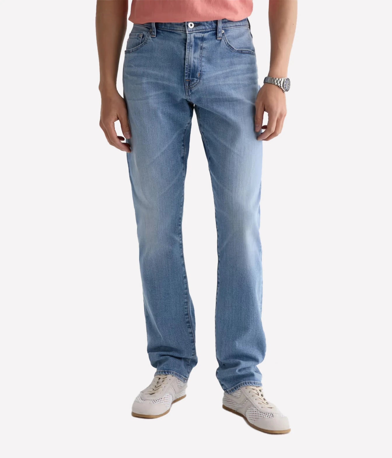 A person wearing light blue slim-fit jeans, a coral pink t-shirt, and off-white sneakers, shown from the waist down against a light gray studio background.