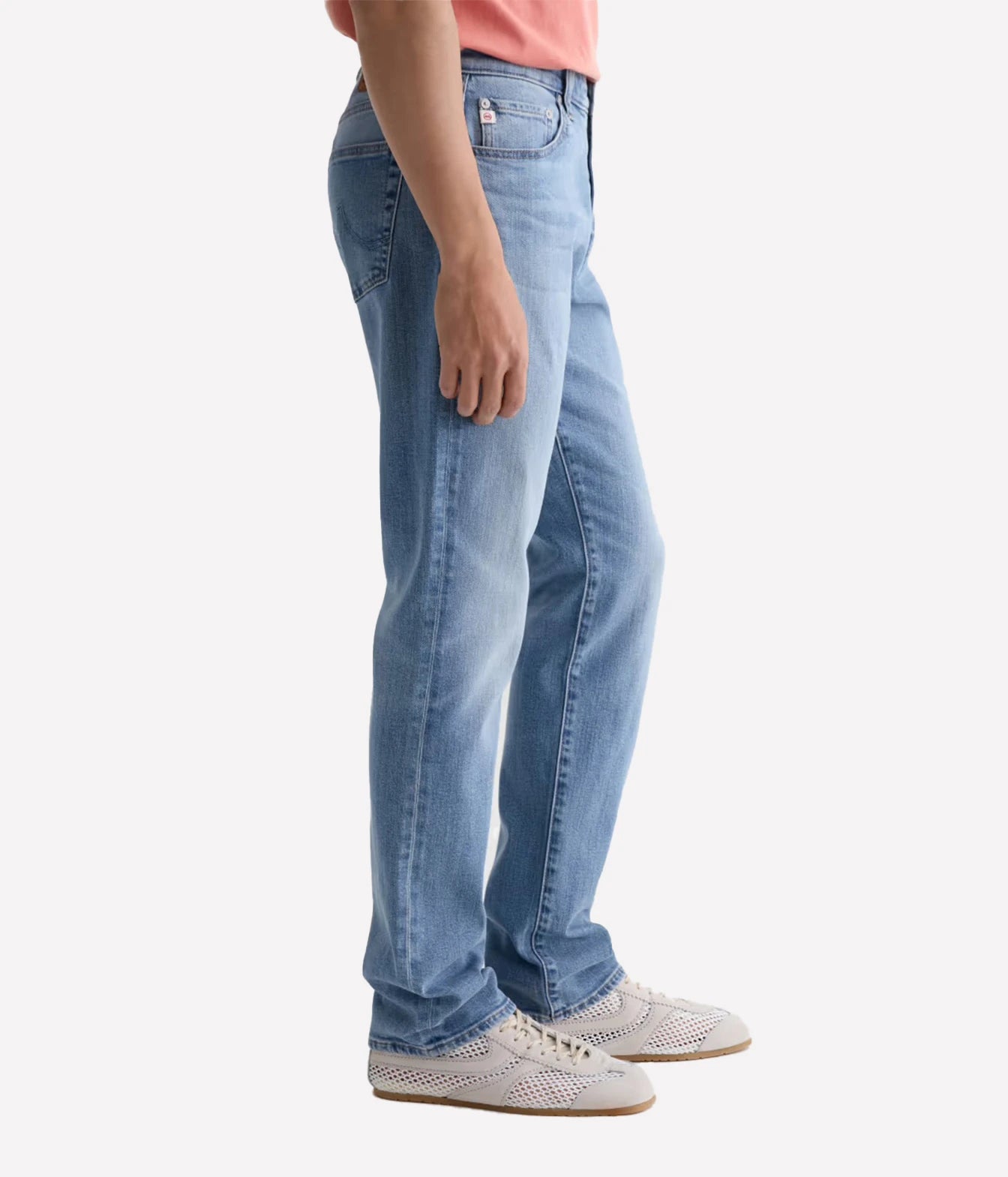 A person shown from the waist down wearing light-wash blue jeans, a coral pink top, and white perforated sneakers against a plain light gray studio background.