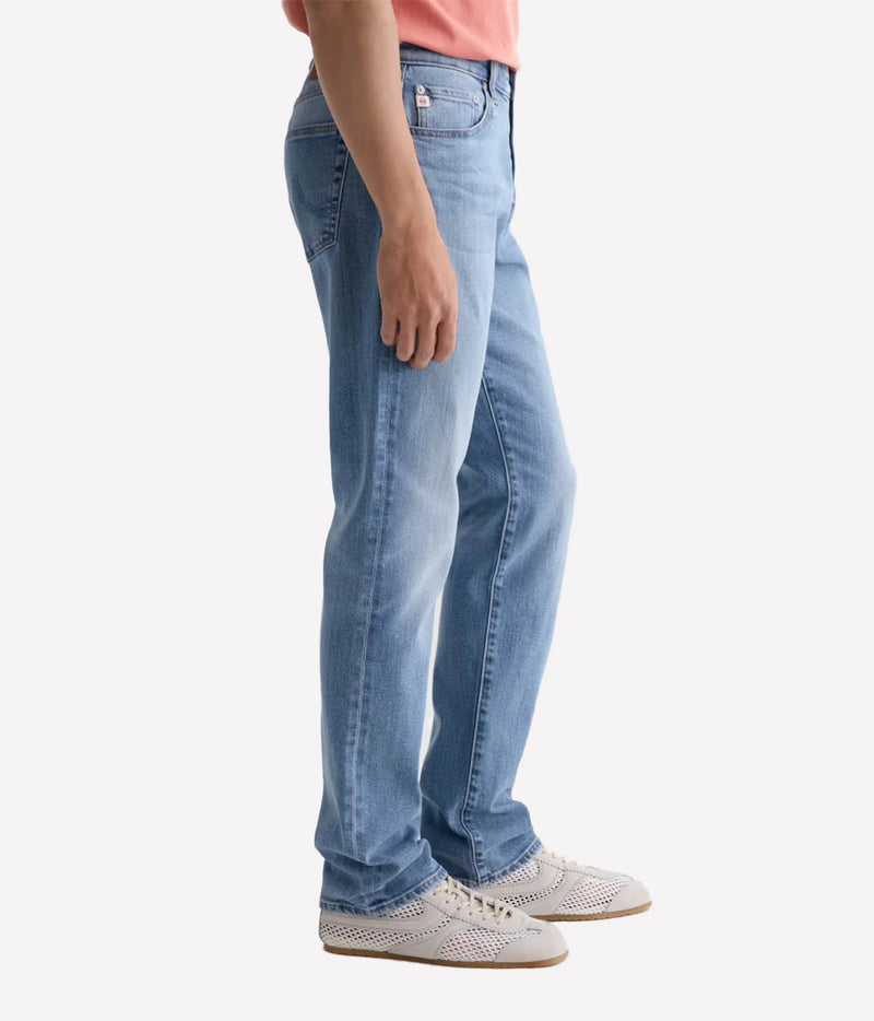 A person shown from the waist down wearing light-wash blue jeans, a coral pink top, and white perforated sneakers against a plain light gray studio background.