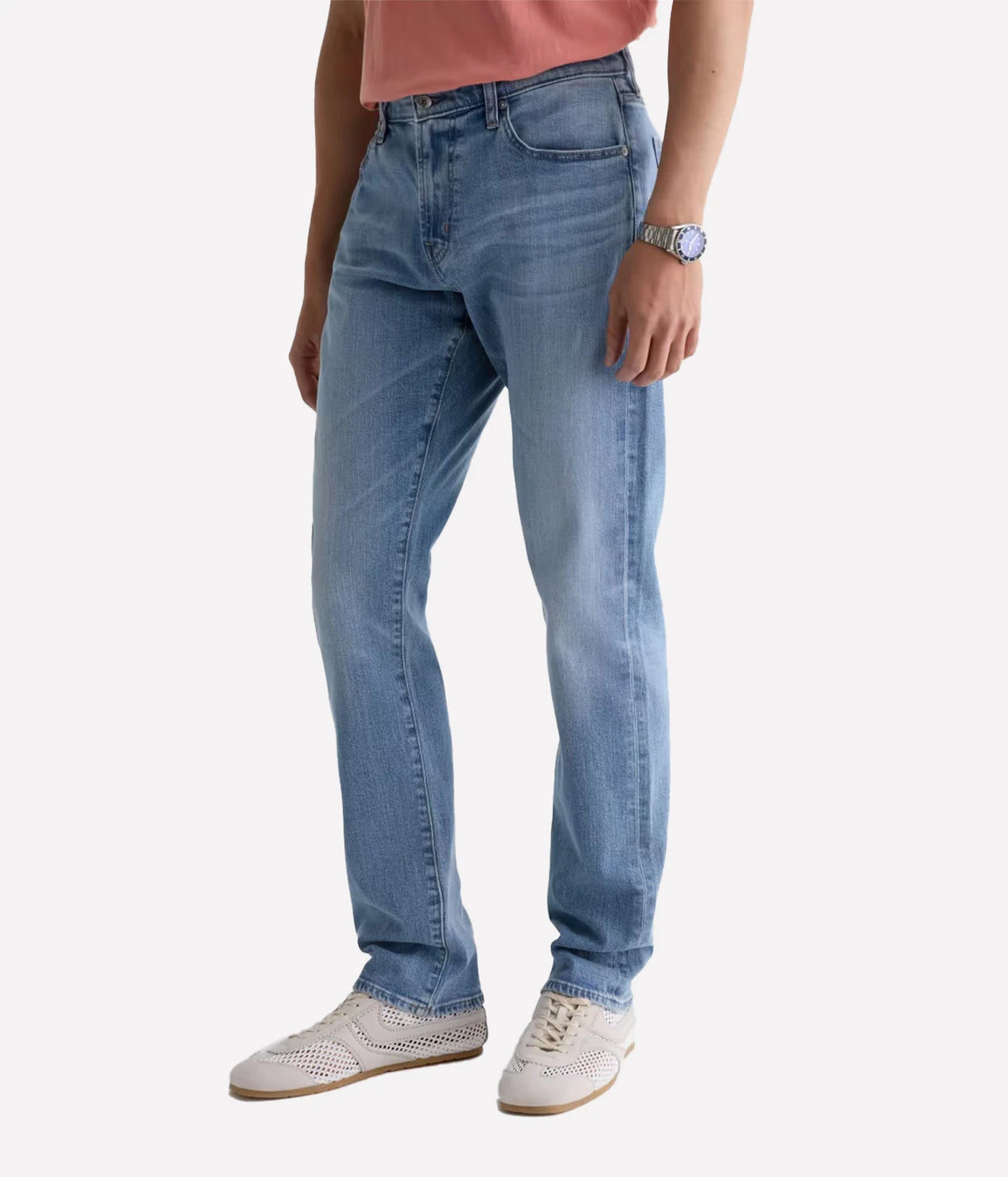 A person wearing light blue slim-fit jeans, a pink top, and white sneakers, shown from the waist down against a light gray studio background.