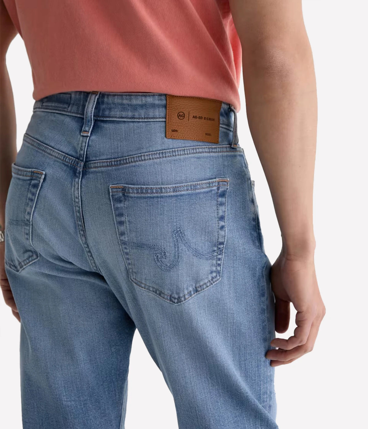Close-up back view of a person wearing light blue denim jeans with a coral pink top, showing the back pocket and leather waistband patch.