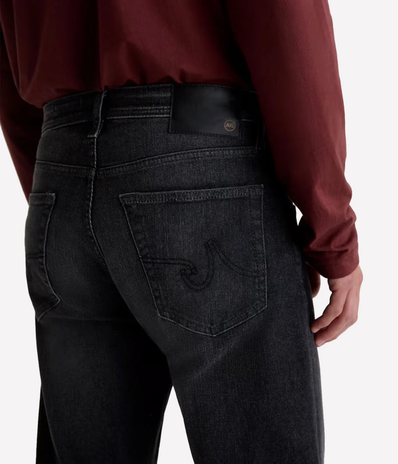 Close-up back view of a person wearing dark charcoal gray denim jeans with a black leather waistband patch and a curved embroidered logo on the back pocket, paired with a burgundy long-sleeve shirt against a light gray background.