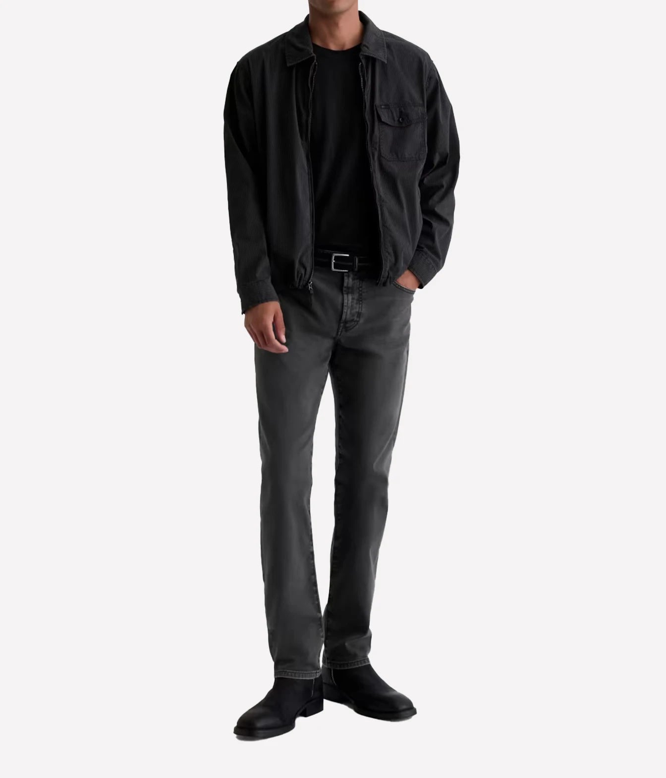 A man wearing a black overshirt, black t-shirt, dark gray slim-fit jeans, and black boots, standing against a light gray background.