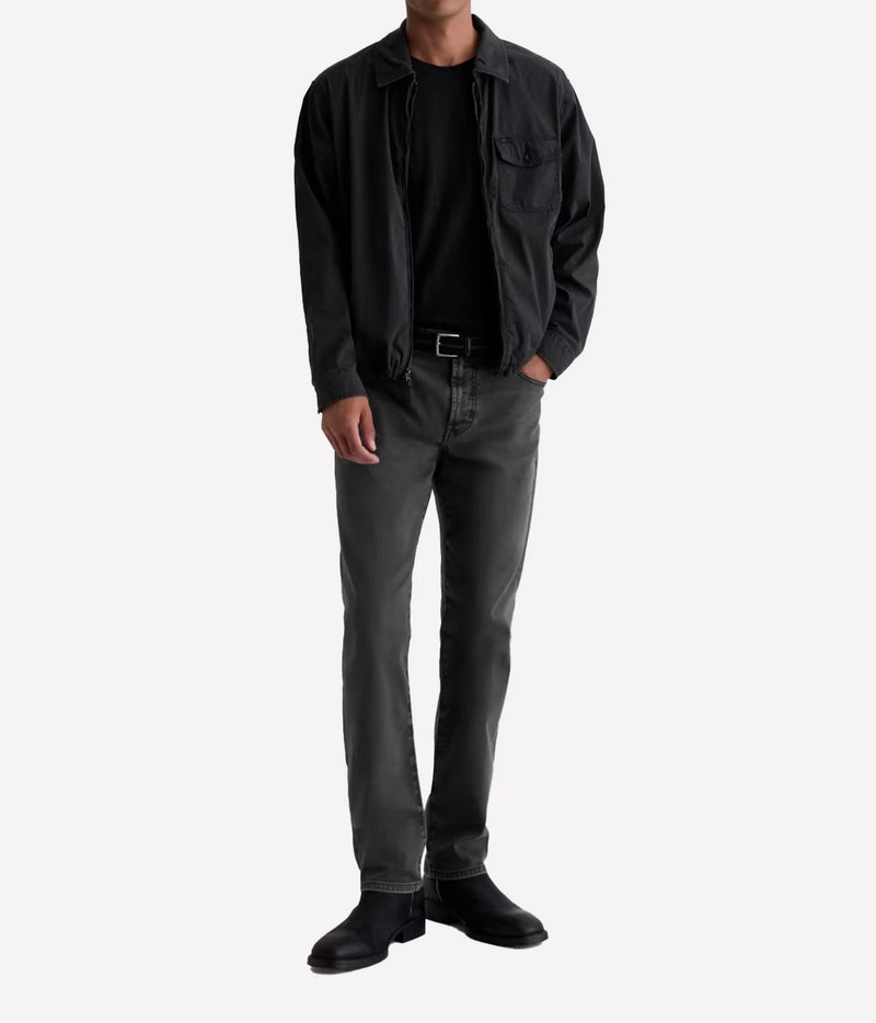 A man wearing a black overshirt, black t-shirt, dark gray slim-fit jeans, and black boots, standing against a light gray background.