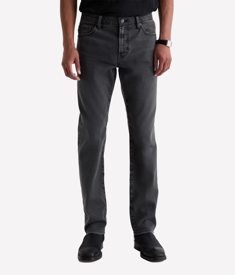 A person wearing slim-fit dark gray washed jeans, a black shirt, and black boots, standing against a light gray studio background.