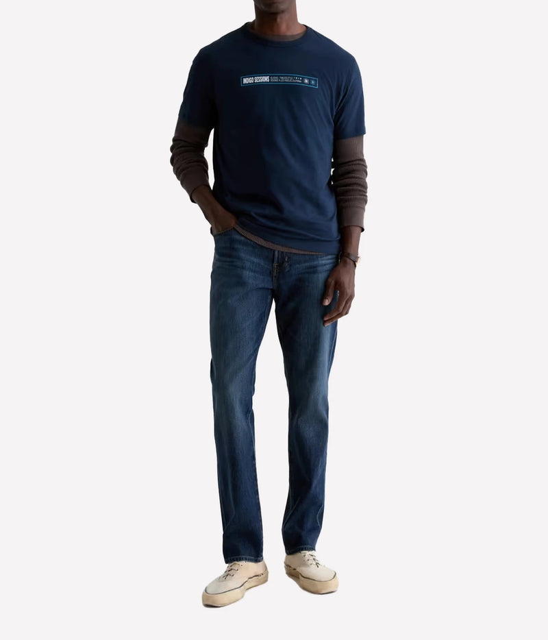 A man wearing a navy blue long-sleeve t-shirt with brown sleeves, dark blue jeans, and off-white sneakers, standing against a light gray background.