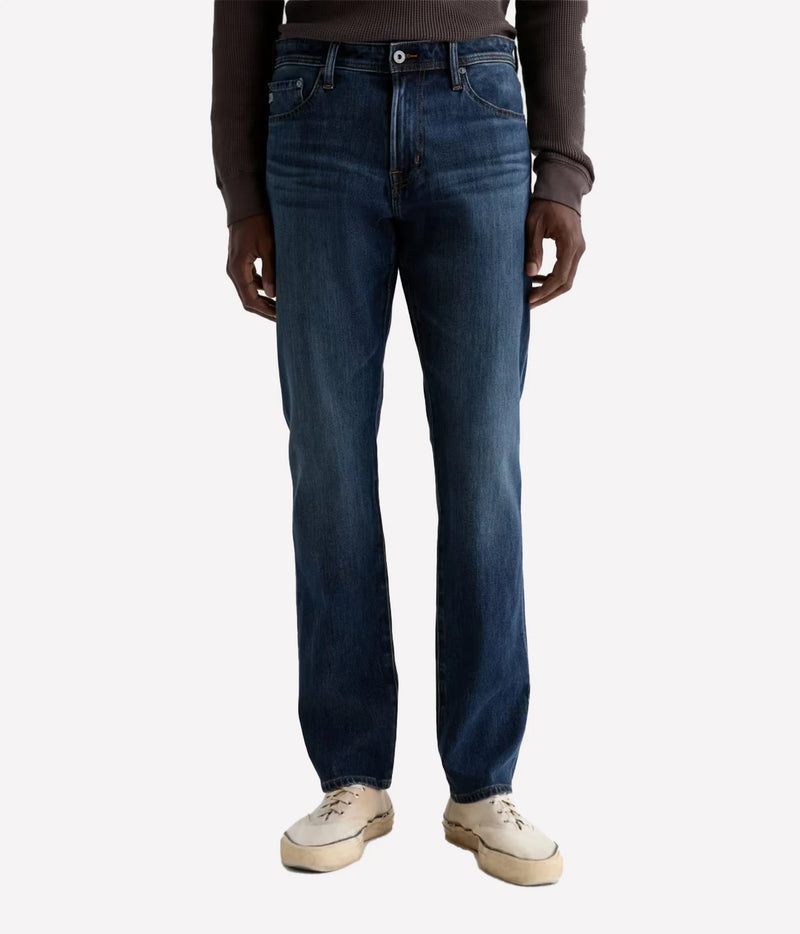 A person wearing dark blue straight-leg denim jeans, a dark brown long-sleeve top, and off-white sneakers, shown from the waist down against a light gray background.