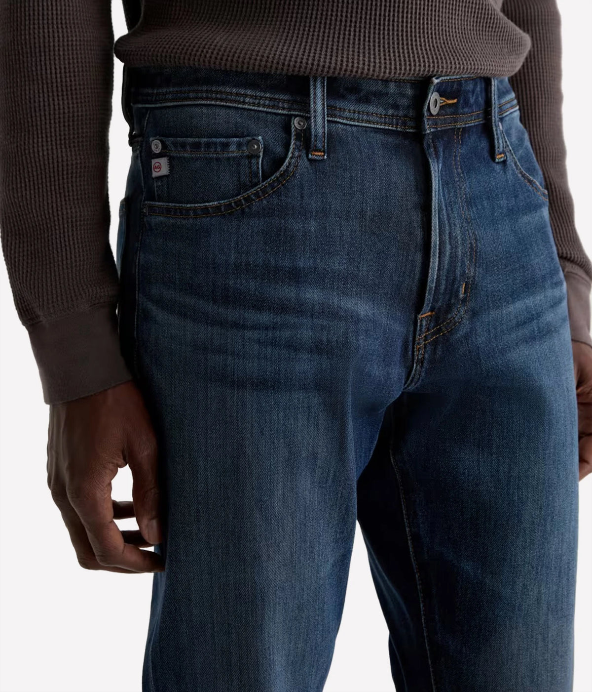 Close-up of a person wearing dark blue denim jeans and a brown ribbed sweater, showing the front and side of the jeans with a small red and white brand tab on the pocket.