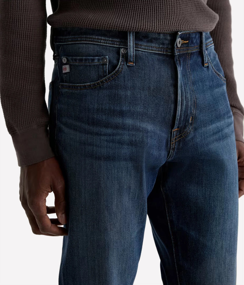 Close-up of a person wearing dark blue denim jeans and a brown ribbed sweater, showing the front and side of the jeans with a small red and white brand tab on the pocket.