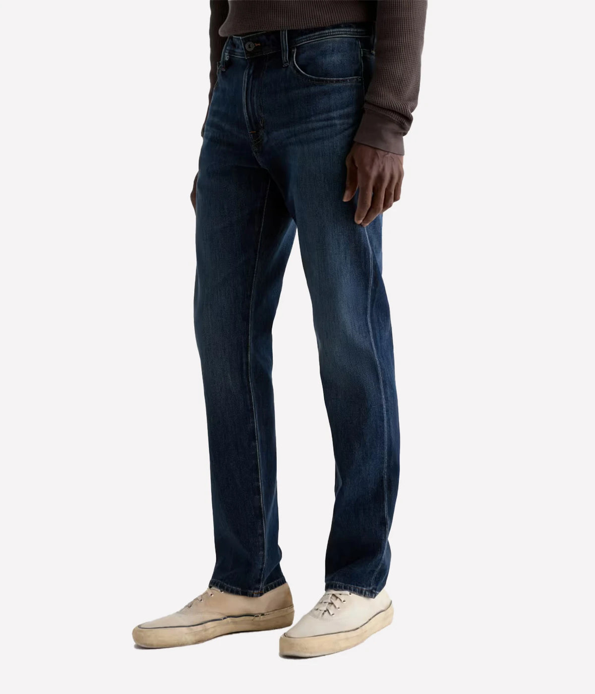 A person wearing dark blue straight-leg denim jeans, a brown long-sleeve top, and beige sneakers, shown from the waist down against a light gray studio background.