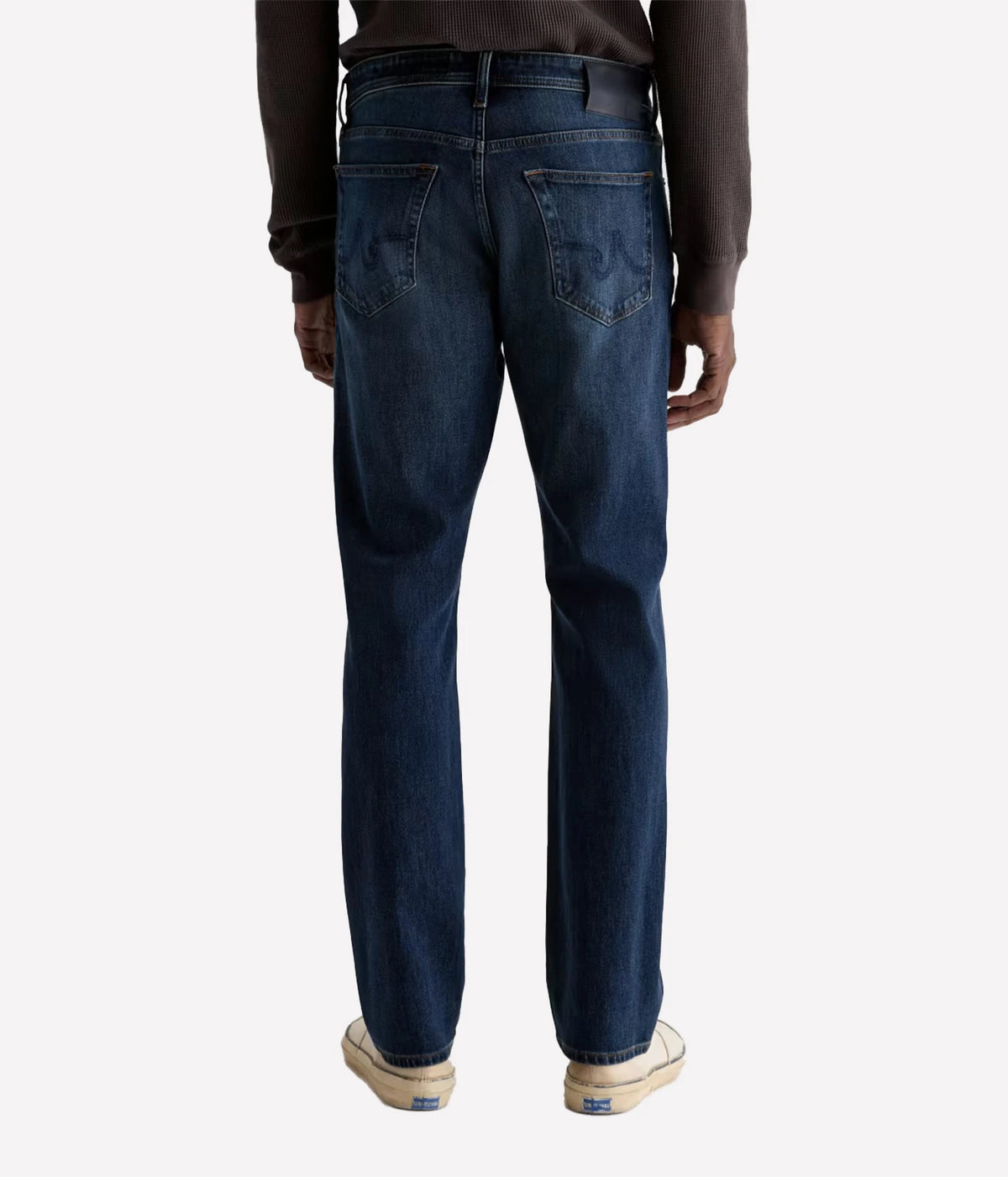Rear view of a person wearing dark blue denim jeans with a brown long-sleeve top and white sneakers, shown against a light gray studio background.