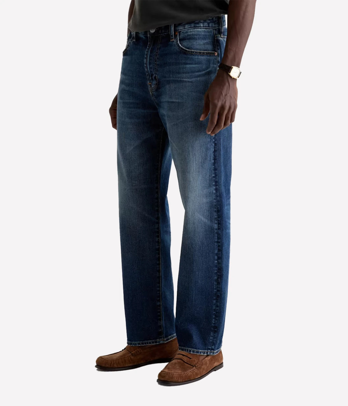 A person wearing dark blue straight-leg denim jeans, brown suede loafers, and a black shirt, shown from the waist down against a light gray studio background.