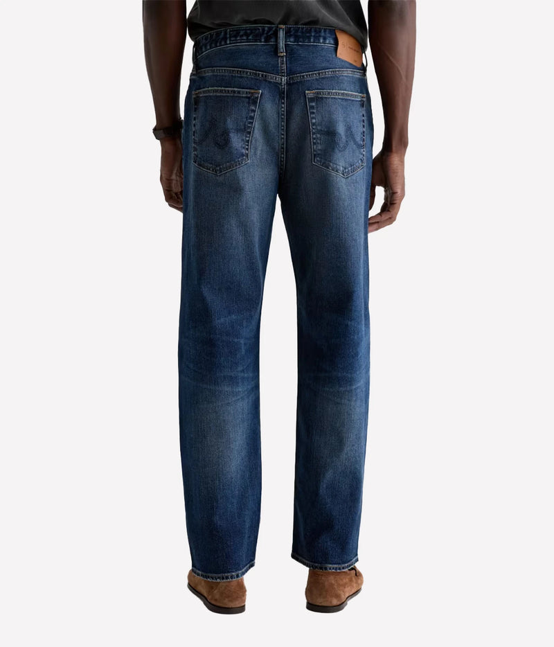 Rear view of a person wearing blue denim jeans with a dark top and brown shoes, shown against a light gray studio background.
