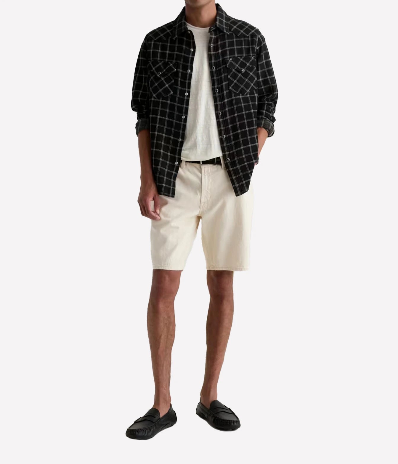 A man wearing a black and white plaid button-down shirt over a white t-shirt, paired with off-white shorts and black loafers, photographed against a solid light gray background.