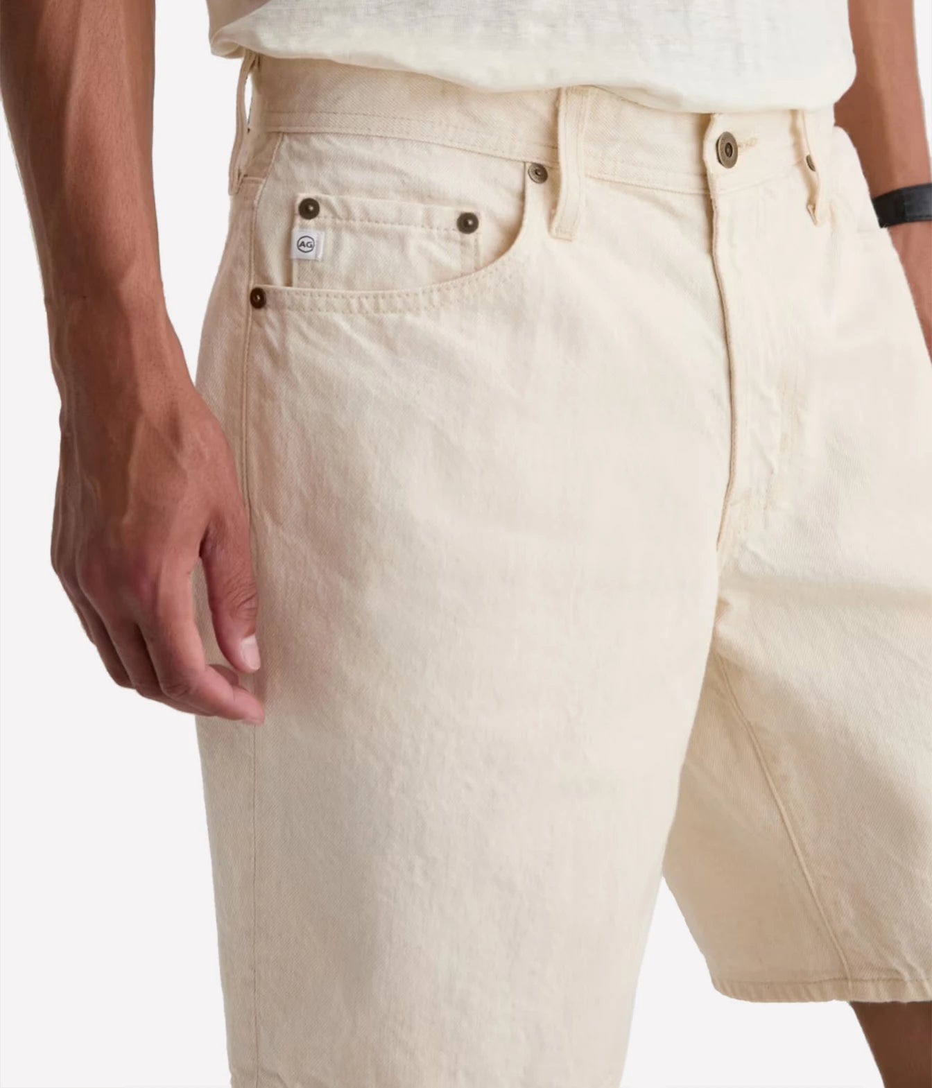 Close-up of a person wearing light cream or off-white denim shorts, showing the front pocket, belt loops, waistband, and part of the hand and torso against a plain light background.