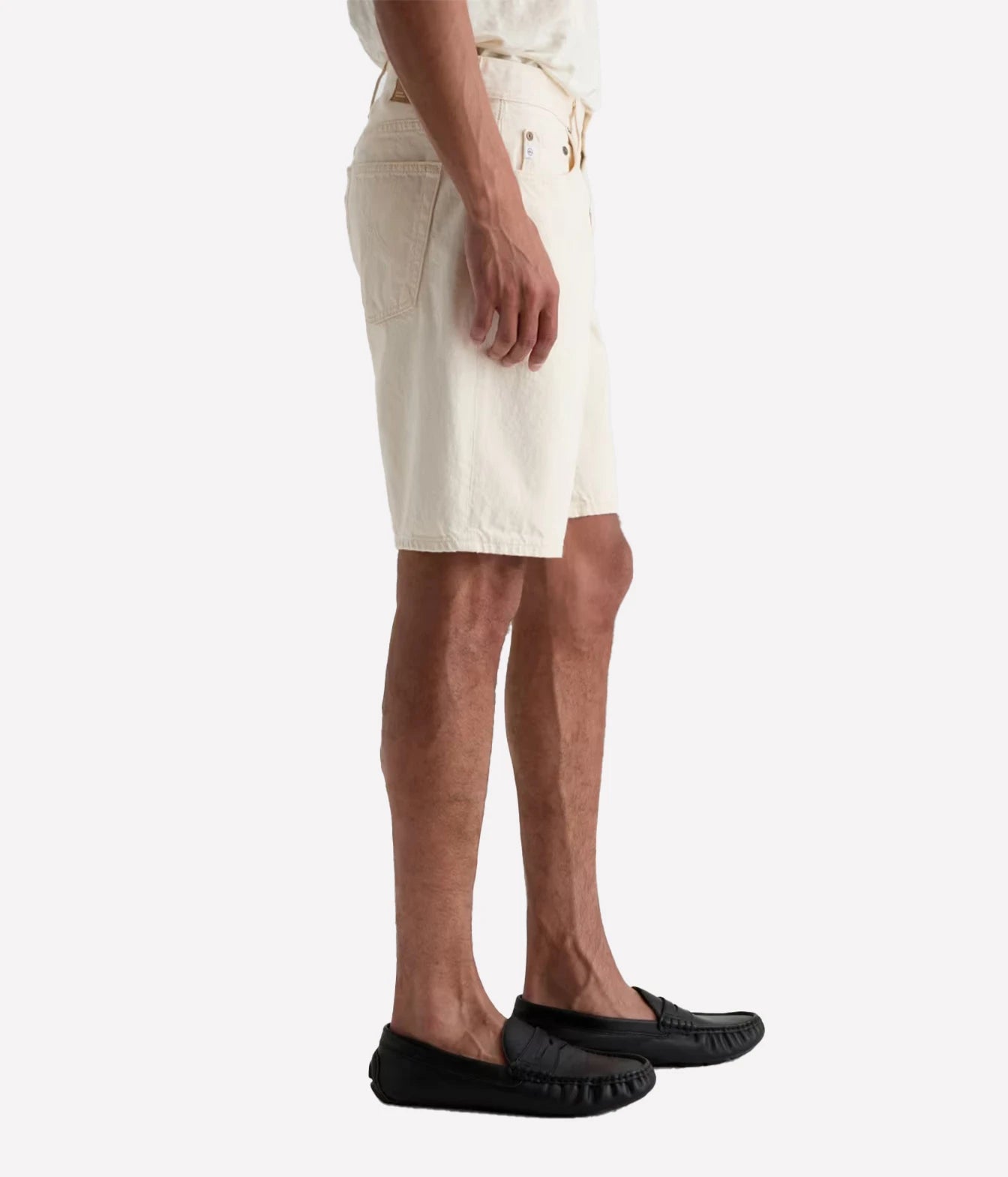 A side profile of a person wearing off-white canvas shorts, a matching off-white shirt, and black leather moccasin loafers, set against a plain light gray background.