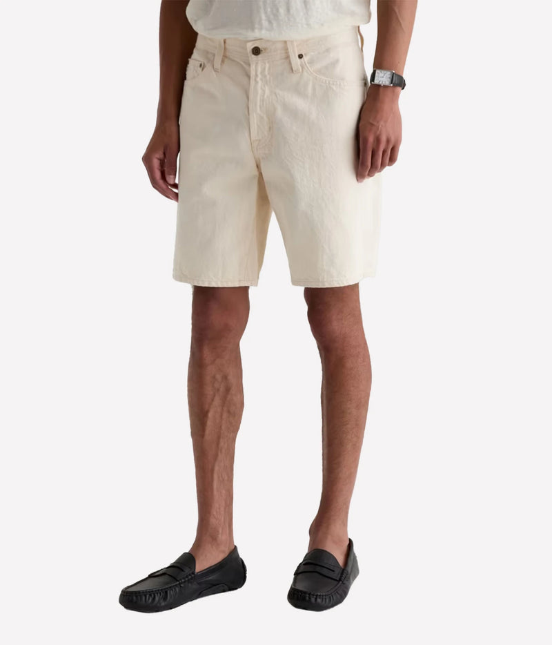 A man wearing off-white textured denim shorts, a white t-shirt, and black leather loafers, shown from the waist down against a light gray studio background.