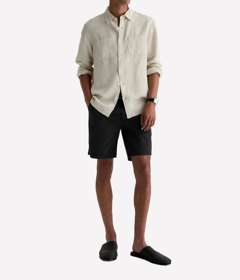A man wearing a light cream linen button-down shirt, black shorts, and black slip-on shoes, standing against a plain white background.