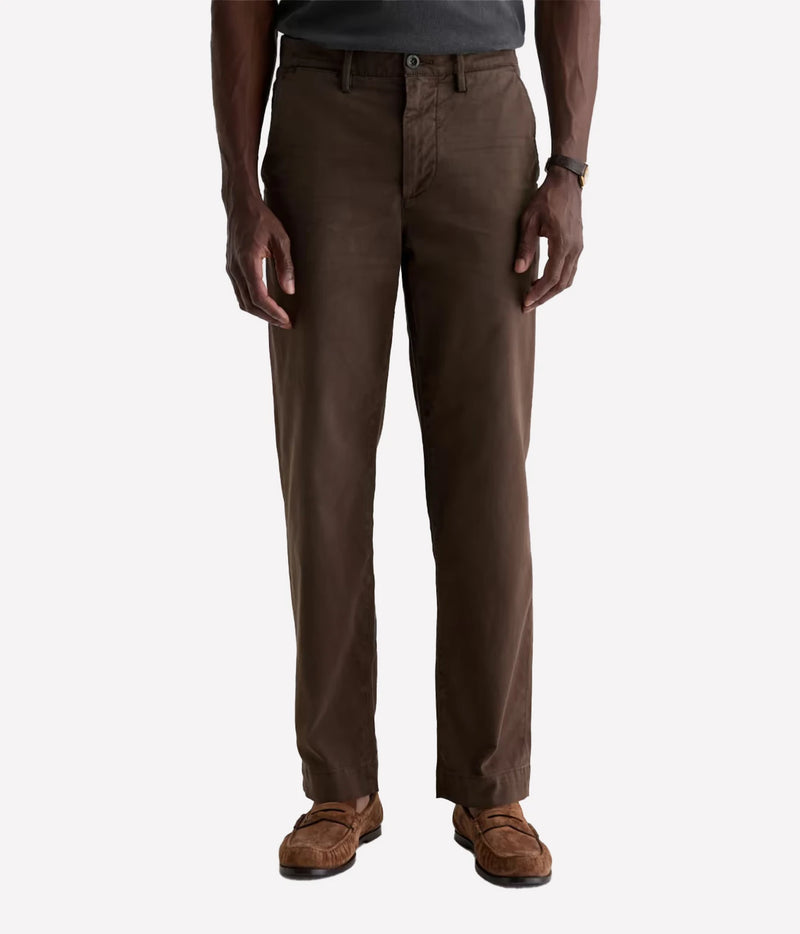 A man wearing brown slim-fit chinos with a dark gray t-shirt and brown suede loafers, shown from the waist down on a light gray background.