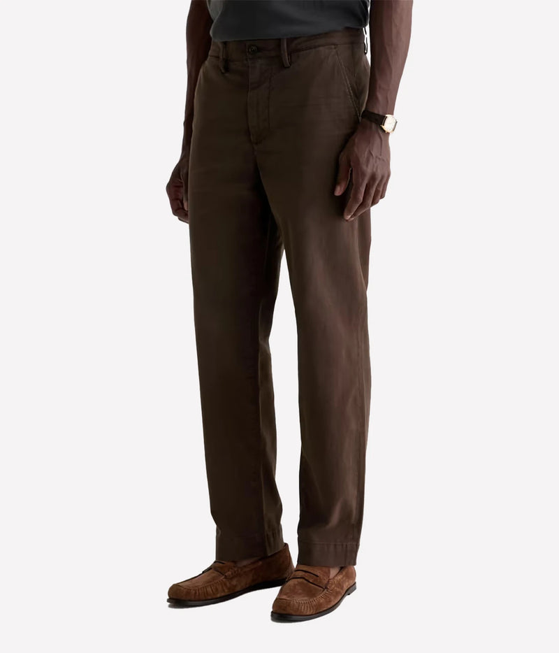A man wearing dark brown chino pants, a dark gray shirt, and brown suede loafers, shown from the waist down on a light gray background.
