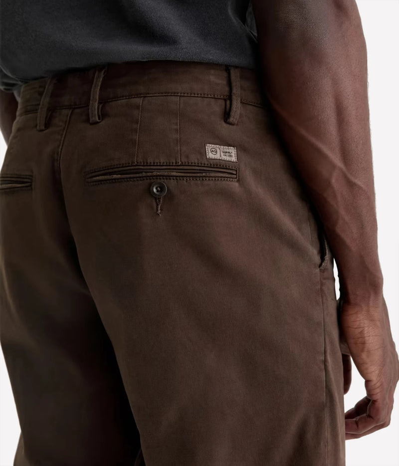 Close-up back view of a person wearing dark brown chino pants, showing the waistband, belt loops, back pockets, and a small rectangular brand label on the upper right side of the pants.