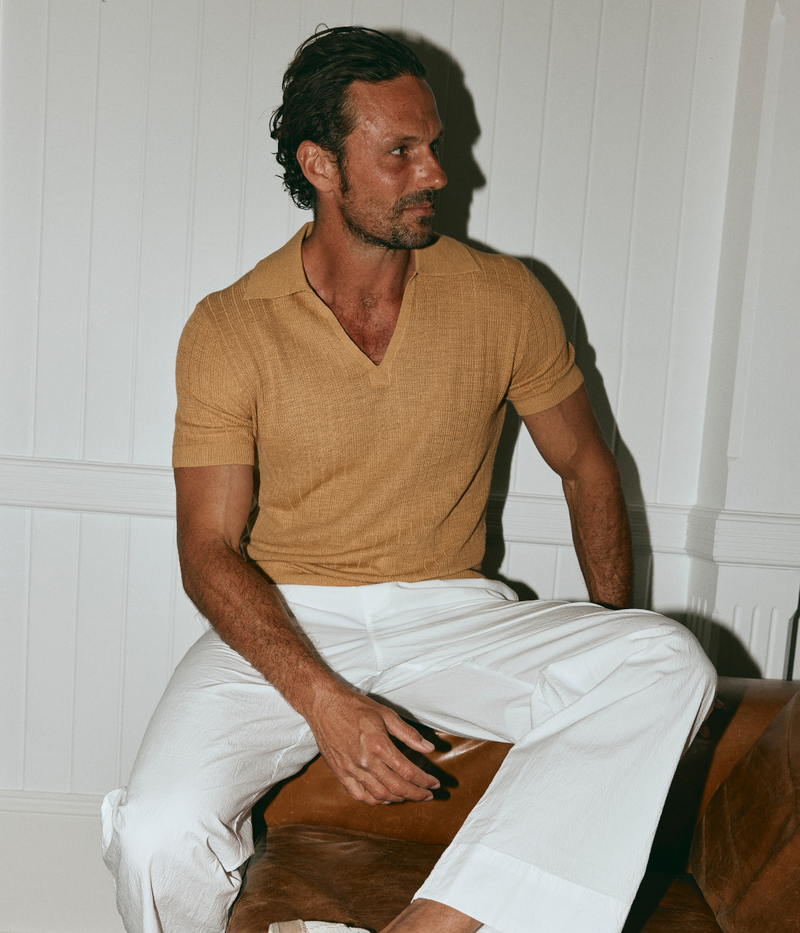 Man sitting on a wooden stool wearing a mustard yellow shirt and white pants against a white paneled wall.