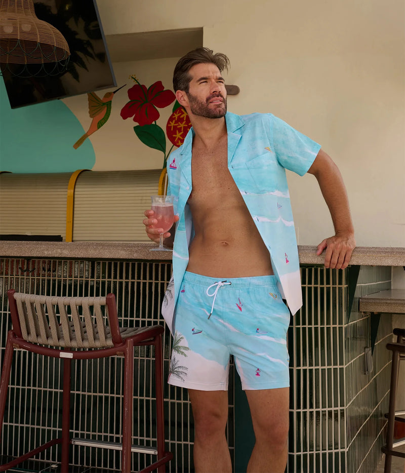 A man wearing light blue tropical print swim trunks and a matching open short-sleeve shirt stands at a bar counter holding a pink drink.