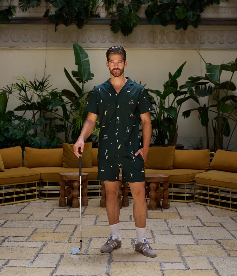 A man stands in a tropical patio lounge setting wearing a dark green short-sleeve button-up shirt and matching shorts with a white tropical print. He is holding a golf club and wearing white socks and brown shoes.