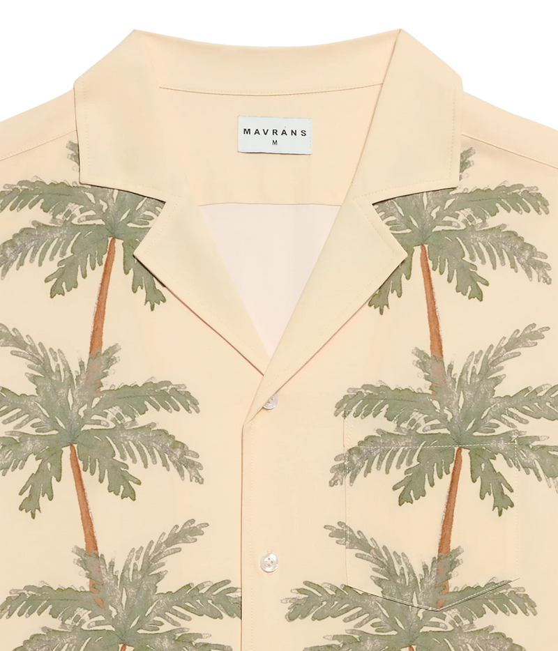 A close-up of a Mavrans men's short-sleeve button-up shirt in a light cream color, featuring a green palm tree print and a white brand label at the collar.