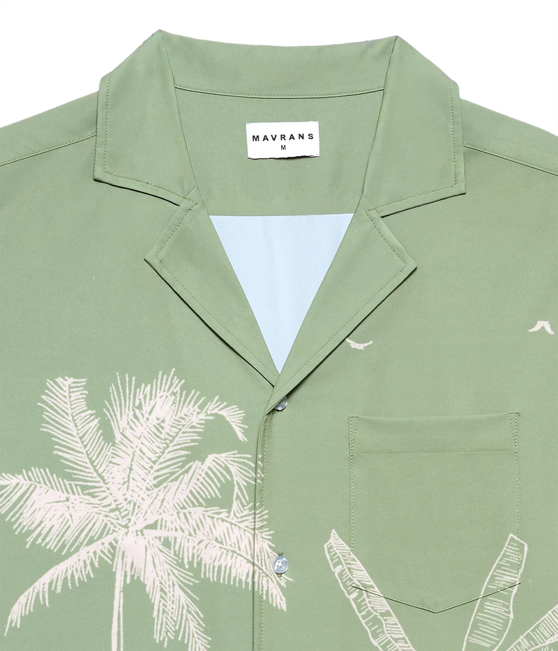 A close-up of a sage green short-sleeve camp collar shirt by Mavrans, featuring a white palm tree print on the front, a white inner collar lining, and a white brand label.
